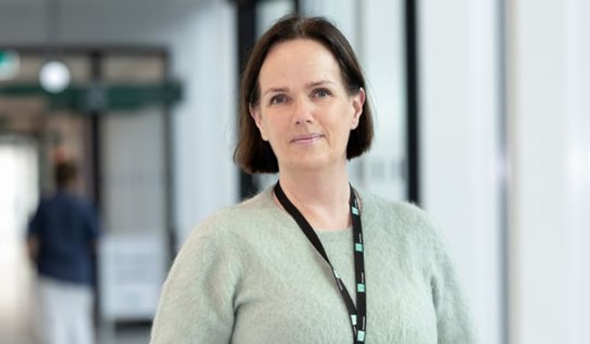 En kvinna står i en korridor med en namnbricka på halsen. Grön tröja i en sjukhusgång.